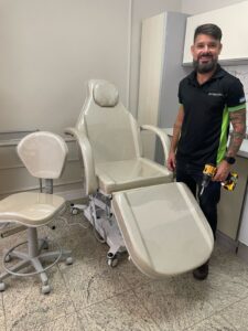 Trabalhador uniformizado da FR Montagens sorri ao lado de cadeira de dentista após montagem de móvel